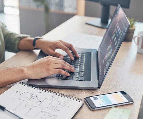 Software developer writing custom code on a laptop with colorful syntax highlighting, with a mobile app wireframe and notebook on a wooden desk